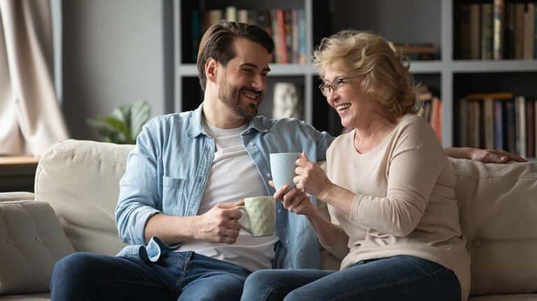 Emotional happy young handsome man telling funny joke or story to laughing middle aged 60s mother, relaxing on comfortable couch with cups of tea, family communication trustful relations concept | 8 Proven Ways To Effectively Communicate With Your Aging Parents