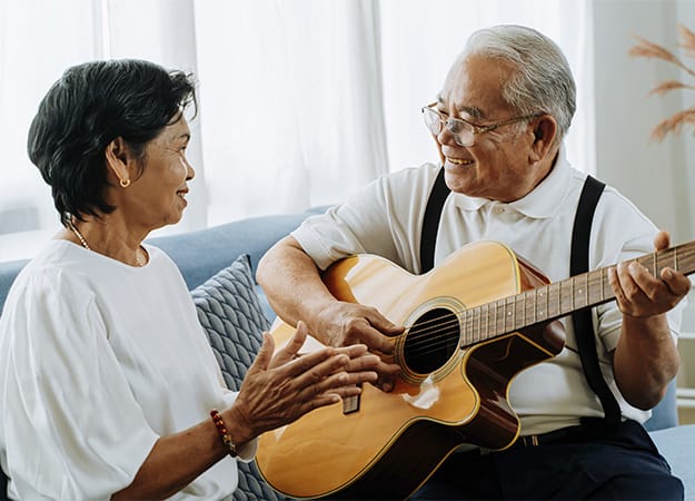 12Oaks-Asian senior couple sitting on the sofa and playing acoustic guitar together-as- 6.Try Music Activities