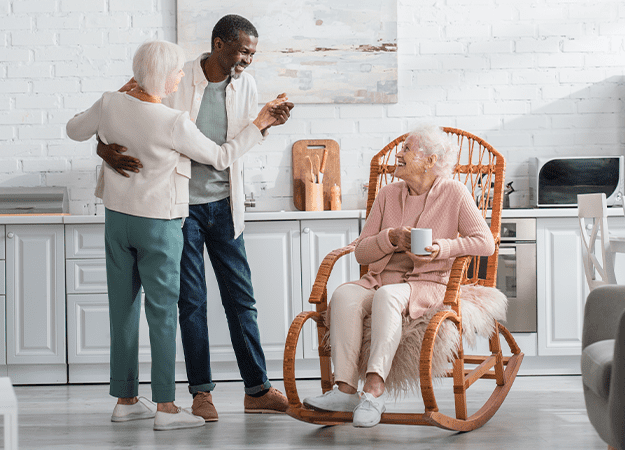 12Oaks-Smiling senior woman holding cup in rocking chair near interracial friends dancing in nursing home-ss-Benefits of Music Therapy for Alzheimer’s