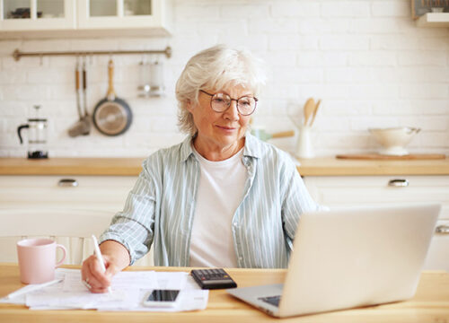 12Oaks-Senior mature female in glasses sitting at kitchen counter in front of laptop computer-as-How Much Does Assisted Living Cost_