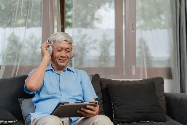 Asian senior man relax at home. Asian older male happy wear headphone using tablet listening podcast while lying on sofa in living room at home concept