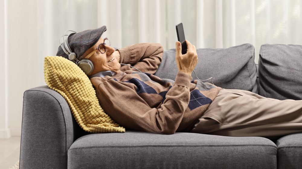 Elderly man on a sofa holding a mobile phone and listening to music on headphones