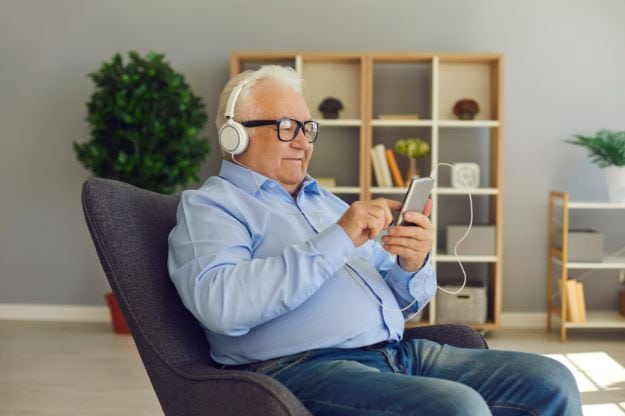 Happy senior man listening to favorite music or podcast wearing headphones and using smartphone