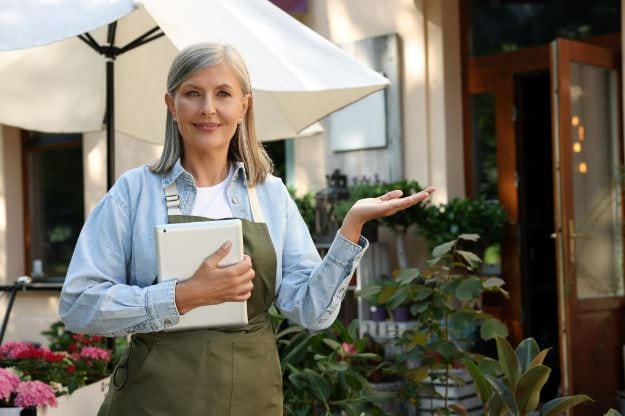 Happy business owner with tablet inviting to come into her flower shop outdoors
