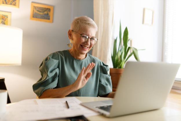 Senior trendy elegant female teacher in glasses with stylish haircut waving hand at camera of laptop saluting her students in beginning of online lesson