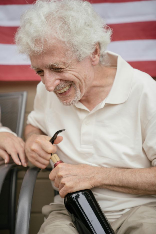 Cheerful Senior Man Sitting in Chair and Opening Wine Bottle 